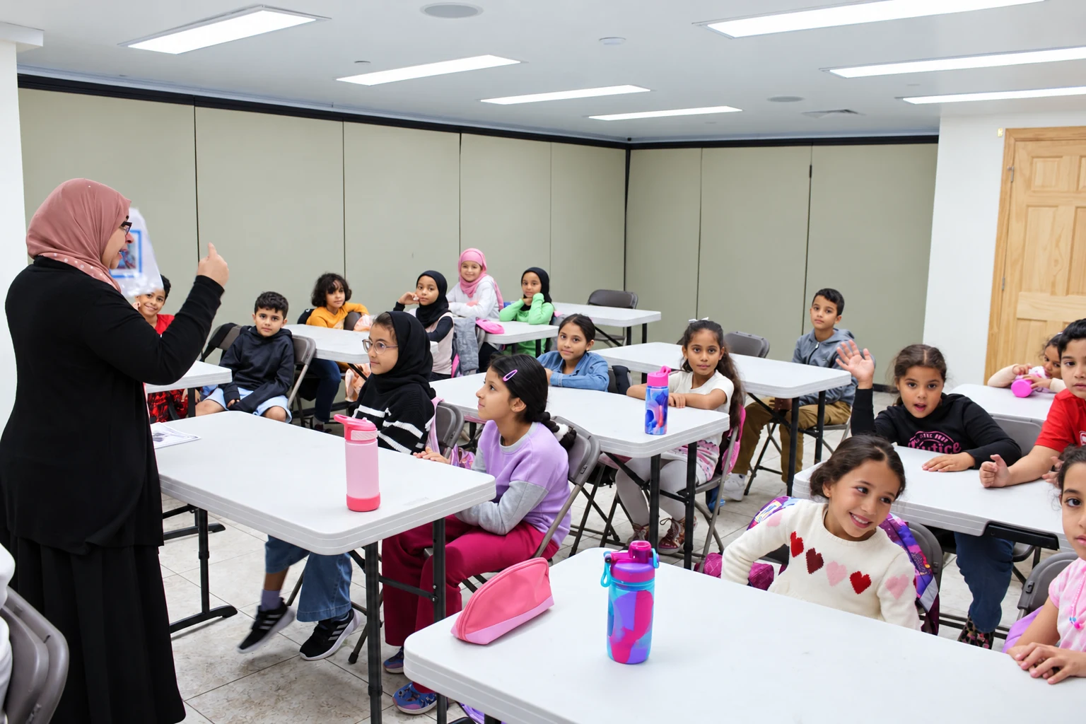 An Al Huda teacher leading a classroom lesson while students listen and raise their hands.